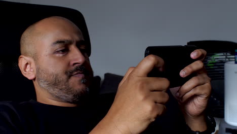 Bald-UK-Asian-Bangladeshi-Man-sits-comfortably-in-a-rolling-chair,-tapping-away-on-his-mobile-phone-screen,-illustrating-the-idea-of-leisure-and-entertainment-at-home