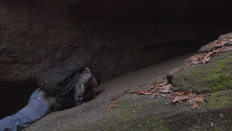 a-bearded-hiker-climbs-up-a-steep-rocky-ledge-pulling-himself-to-his-feet-and-entering-a-dark-cervice-in-the-rocky-moss-covered-formations