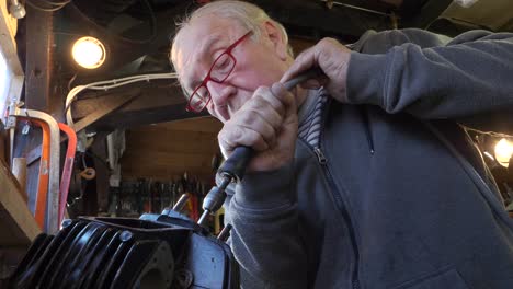 Senior-craftsman-uses-honing-tool-to-shape-engine-block-in-small-dark-workshop