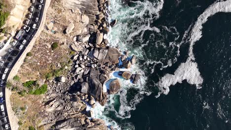 Drone-De-Arriba-Hacia-Abajo-De-Una-Carretera-Muy-Transitada-En-Ciudad-Del-Cabo,-Sudáfrica-A-Lo-Largo-De-La-Costa,-Las-Olas-Chocan-Contra-Las-Rocas