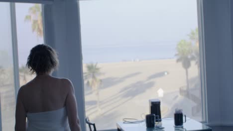 Middle-aged-white-woman-walking-with-white-towel-in-room-and-looking-out-window-of-Pacific-Ocean-in-Venice-Beach,-California-in-slow-motion