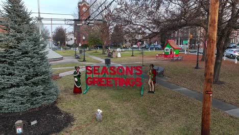 Season-Greetings-Sign-in-park-beside-Christmas-tree-and-church-in-background
