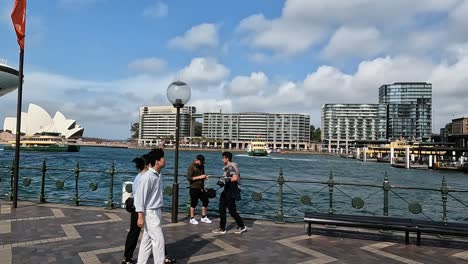 Sydney-harbour-with-ferries,-skyscrapers,-and-a-pedestrian-on-a-sunny-day,-waterfront-promenade-view