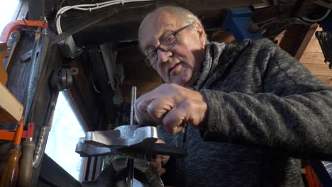 Senior-caucasian-worker-uses-hand-file-to-work-on-a-metal-piece-in-a-small-dark-workshop