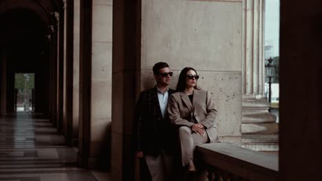 Stilvolles-Paar-Mit-Schwarzer-Sonnenbrille-Lehnt-Und-Sitzt-An-Der-Wand-Zwischen-Den-Bögen-Und-Genießt-Den-Blick-Auf-Den-Leeren-Platz-Rund-Um-Das-Museum-Du-Louvre-Mit-Seinen-Borque-Gebäuden-In-Paris,-Frankreich