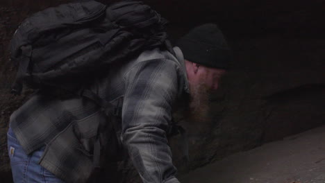 a-hiker-with-a-big-red-beard-and-a-plaid-flannel-shirt-carrying-a-backpack-climbs-his-way-up-a-rocky-ledge-to-reach-a-dark-crevice-in-the-rocky-cavern