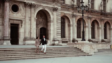 Elegant-couple-in-suits-walking-down-the-strairs-looking-at-each-other-at-the-baroque-royal-residence-buildings-next-to-the-Louve-without-any-tourists-in-Paris-France---wide---modern-classy-lifestyle