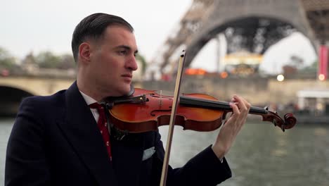 A-caucasian-male-violinist-in-a-black-suit-and-with-a-red-tie-playing-on-his-violin-in-front-of-the-famous-Eiffel-Tower-in-Paris-France-and-the-river-seine---close-up-cinematic