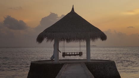 Woman-sitting-down-on-swing-below-thatched-roof-gazebo-during-sunset-at-sea