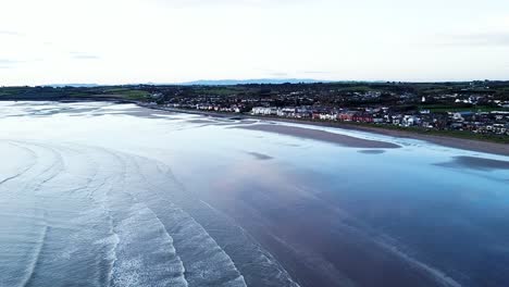 An-establishing-aerial-drone-shot-of-the-scenic-Irish-Sea-coastline-with-calm-waves-rolling-into-the-shore-at-Skerries-Beach-during-sunrise,-Dublin,-Ireland