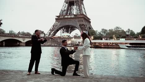 Un-Hombre-De-Traje-Negro-Sobre-Sus-Rodillas-Le-Propone-Matrimonio-A-Su-Novia-Con-Un-Elegante-Traje-Blanco-Para-Convertirse-En-Su-Futura-Esposa-Con-Un-Violinista-Junto-A-Ellos-Frente-A-La-Torre-Eiffel-En-París,-Francia,-Y-Confirma-Que-Sí.