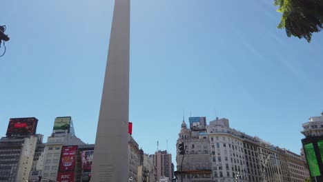 Obelisk-of-Buenos-Aires-City-Argentina-under-Sunshine-Skyline,-Taxis,-Cars-and-Traffic-driving-by-9-de-Julio-Famous-Downtown-Landmark,-Avenue-and-Classical-Buildings