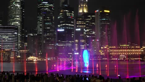 People-gathered-at-the-Marina-bay-sands-event-plaza,-watching-SPECTRA-light-and-water-show-in-the-evening-with-downtown-cityscape-in-the-background,-static-shot-of-Singapore-night-entertainment