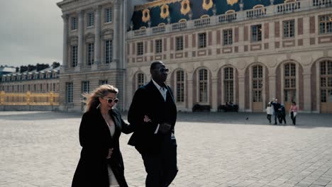 Elegantes-Mischlingspaar-Mit-Sonnenbrille-Läuft-über-Den-Cour-Royale-Zum-Berühmten-Schloss-Versailles-In-Paris,-Frankreich-–-Orbitalaufnahme,-Die-Das-Hauptgebäude-Freigibt