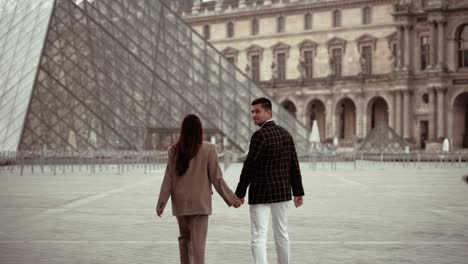 Pareja-Elegante-Caminando-Sobre-Un-Lugar-Vacío-Del-Museo-Del-Louvre-Mirando-Hacia-La-Cámara-Rodeada-Por-Los-Edificios-Barrocos-De-La-Residencia-Real-En-París,-Francia:-Estilo-De-Vida-Moderno-Y-Trajes-Para-Amantes-De-Las-Citas