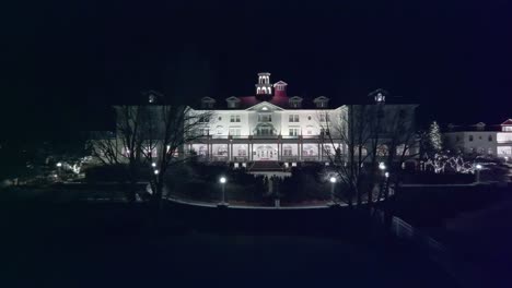 Aerial-frontal-dolly-to-entrance-of-creepy-old-white-hotel-at-night,-fly-between-trees