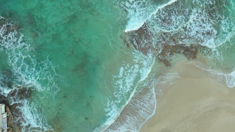 top-down-aerial-shot-capturing-the-gentle-waves-of-the-turquoise-sea-as-they-wash-onto-a-sandy-beach,-creating-a-lacy-pattern-of-foam-along-the-shore