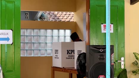 Man-in-traditional-attire-casting-vote-in-Indonesian-election,-ballot-boxes-visible,-indoor-setting