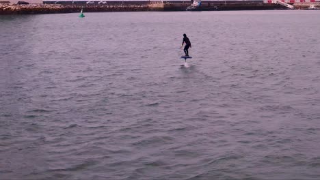Aerial-tracking-of-a-young-man-riding-on-an-electric-hydrofoil-surfboard-on-glassy-blue-ocean-water-with-dock-behind-him