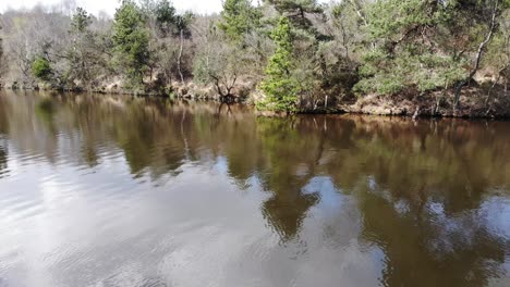 Aerial-Flying-Over-Brown-Water-At-Squabmoor-Reservoir-With-Woodland-Trees-Seen-On-Embankment