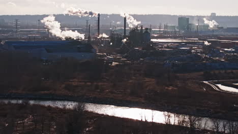 Industrial-complex-on-riverbank-with-smokestacks-emitting-steam,-backlit-by-sunlight,-environmental-impact-concept