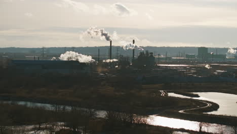 Industrial-complex-with-smokestacks-by-river,-clear-sky,-distant-view