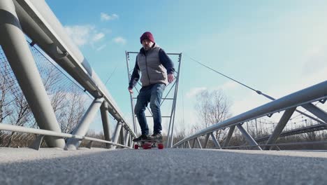 Elderly-man-on-longboard-riding-on-a-pedestrian-bridge-under-a-clear-sky,-wearing-jeans,-a-jacket,-and-a-red-cap