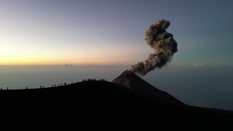 Drone-view-in-Guatemala-flying-over-a-volcano-crater-with-an-erupting-volcano-at-sunrise,-people-on-the-rim-surrounded-by-clouds