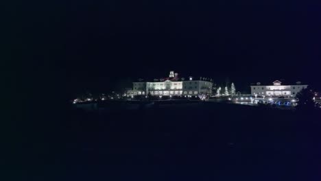 Low-angle-aerial-dolly-establishes-grand-white-hotel-with-parking-lot-at-night-as-spotlight-crosses-grass