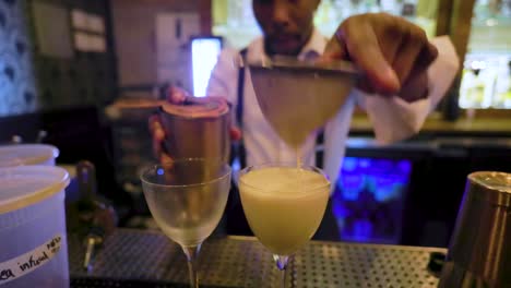 Schwarzer-Männlicher-Barkeeper-Gießt-Einen-Frisch-Zubereiteten-Cocktail-In-Ein-Glas,-Um-Ihn-Einem-Kunden-Einer-Lokalen-Bar-Zu-Servieren