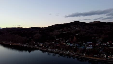 Twilight-descends-on-a-tranquil-riverside-town-with-hills,-aerial-view