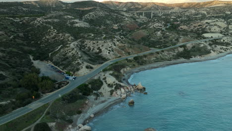 Aerial-view-of-a-coastal-road-near-Aphrodite's-Rock-in-Cyprus,-winding-along-the-shoreline-and-hilly-terrain