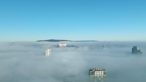 A-sunlit-aerial-journey-as-a-drone-glides-backward,-capturing-the-Bratislava-castle-and-buildings-emerging-through-dense-inversion-clouds