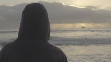 Rear-view-carefree-hooded-young-adult-male-watching-ship-on-the-distant-sunset-horizon