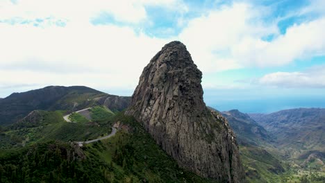 Drone-View-of-Roque-de-Agando-Surrounded-by-Lush-Vegetation,-Curvy-Road,-La-Gomera,-Canary-Islands,-Spain