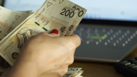Female-hands-counting-200-Polish-zloty-bills-with-a-laptop-in-the-background---house-budget-concept,-close-up-shot