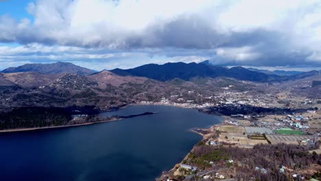 A-serene-lake-with-nearby-town-surrounded-by-mountains-under-a-cloudy-sky,-daytime,-aerial-view