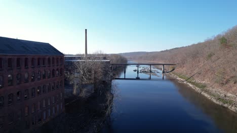 Flying-along-millhouse-on-a-river-with-morning-light-shining-through-its-windows-and-branching-trees-over-the-water-with-a-chimney-and-bridge-in-the-background