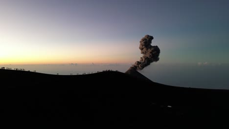 Drohnenansicht-In-Guatemala,-Die-über-Einen-Vulkankrater-Fliegt,-Mit-Einem-Ausbrechenden-Vulkan-Bei-Sonnenaufgang-Und-Schatten-Von-Menschen-Auf-Dem-Rand