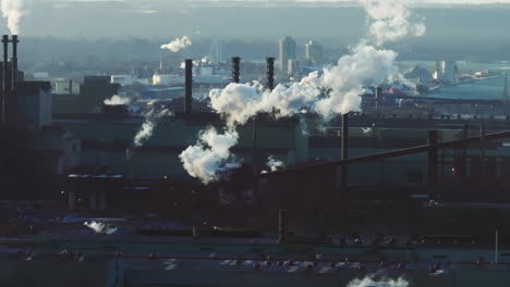 Industrial-complex-with-smokestacks-emitting-steam-and-smoke,-city-backdrop,-early-morning-light