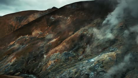 Aerial-drone-tilt-shot-of-a-series-of-steaming-geysers-and-hot-springs-in-a-canyon-amid-snow-capped-mountains-and-hills