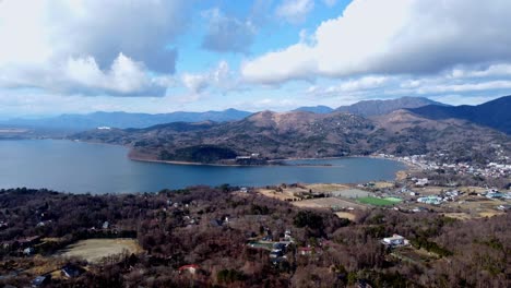 A-serene-lake-beside-a-small-town-with-mountains-under-a-cloudy-sky,-daylight,-aerial-view