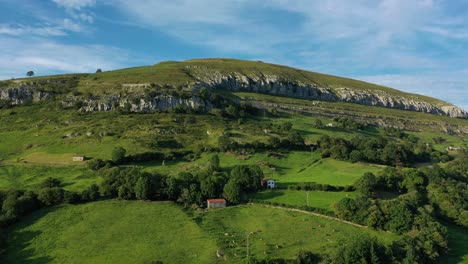 side-flight-seeing-a-white-limestone-mountain-with-green-meadows-and-farm-houses-on-their-farms,-many-separated-with-oak-barriers-on-a-summer-day-with-a-blue-sky-with-clouds-in-Cantabria-Spain
