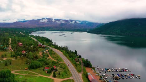 Vista-Aérea-Por-Drones-Del-Hermoso-Lago-Con-Las-Montañas-Rocosas-Al-Fondo-Mientras-Las-Nubes-Cruzan-Los-Picos-Y-Los-Autos-Conducen-Por-La-Carretera-De-Colorado-A-Lo-Largo-De-La-Costa-De-La-Montaña-De-Sombra-Y-El-Gran-Lago