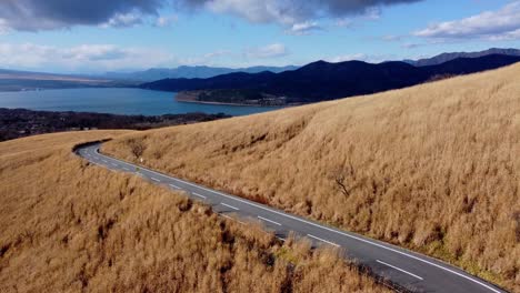 Winding-road-cutting-through-golden-hills-with-a-view-of-a-lake-and-mountains,-under-a-partly-cloudy-sky,-aerial-shot