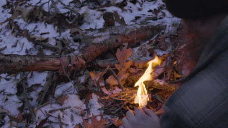 slo-motion-shot-of-a-bearded-man-builds-a-small-campfire-in-the-forest-by-adding-small-twigs-to-feed-the-fire-with-fallen-leaves-and-snow-covering-the-forest-floor