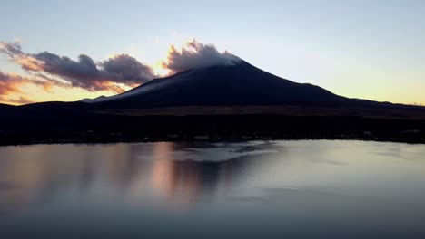 Sunset-hues-reflecting-on-water-with-Mount-Fuji-silhouetted-against-a-dusky-sky,-clouds-gathering