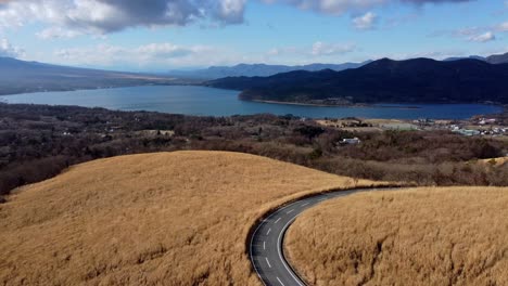 A-curvy-road-cutting-through-golden-grass-with-a-lake-and-mountains-in-the-distance,-aerial-view