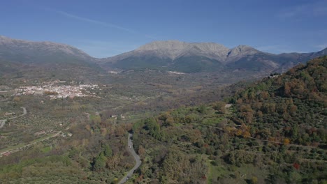 Vuelo-En-Un-Valle-En-Otoño-Con-árboles-Coloridos-Con-Una-Carretera-Y-Autos-Conduciendo,-En-Bosques-De-Pinos-Con-Un-Fondo-De-Montañas-De-Granito-Y-Una-Ciudad-En-Un-Día-Con-Cielo-Azul-En-ávila-españa