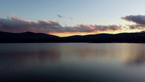 Sunset-over-calm-lake-with-silhouette-of-mountains-and-clouds-reflecting-on-water,-aerial-view